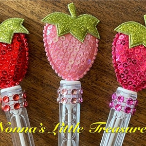 May include: Three clear plastic tubes with a sparkly strawberry-shaped topper. Each strawberry is made of sequins and has a green glitter leaf. The tubes have a band of sparkly rhinestones around the middle.