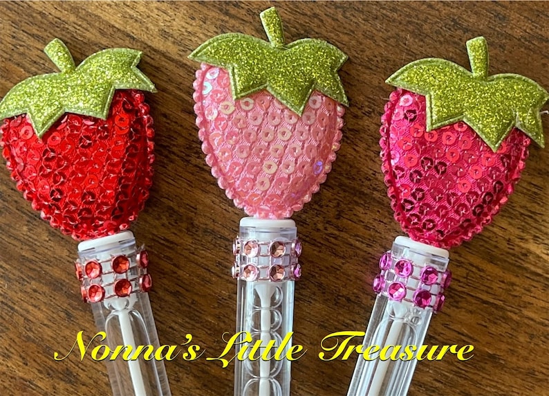 May include: Three clear plastic tubes with a sparkly strawberry-shaped topper. Each strawberry is made of sequins and has a green glitter leaf. The tubes have a band of sparkly rhinestones around the middle.