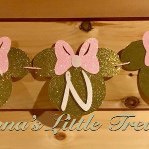May include: A pink and gold glitter banner with the word "ONE" in gold letters and pink bows. The banner is decorated with pink and gold glitter mouse ears. The banner says "Nonna's Little Treasure" at the bottom.