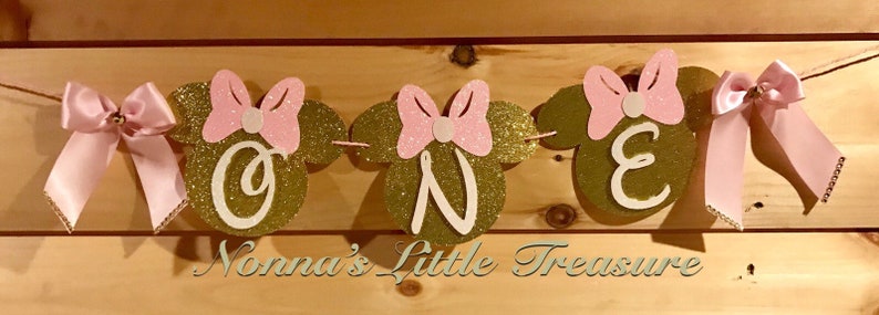 May include: A pink and gold glitter banner with the word "ONE" in gold letters and pink bows. The banner is decorated with pink and gold glitter mouse ears. The banner says "Nonna's Little Treasure" at the bottom.