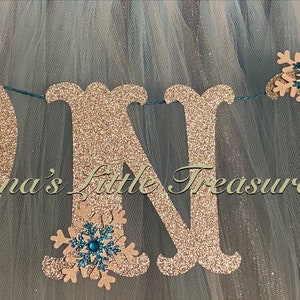 May include: A gold glitter "ONE" banner with teal snowflake accents. The banner is strung together with twine and is perfect for a first birthday party.