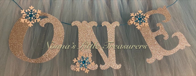 May include: A gold glitter "ONE" banner with teal snowflake accents. The banner is strung together with twine and is perfect for a first birthday party.