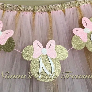 May include: Gold glitter Minnie Mouse head banner with pink bows and white letters spelling out "ONE".