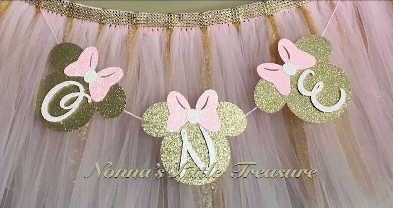 May include: Gold glitter Minnie Mouse head banner with pink bows and white letters spelling out "ONE".