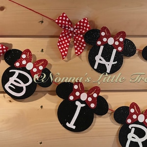 May include: A festive banner with black Mickey Mouse head cutouts spelling out "BIRTHDAY" in white letters. Each cutout has a red bow with white polka dots. The banner is strung on a red string, perfect for a birthday party.