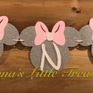 May include: A silver glitter banner with pink bows and the word "ONE" in white letters. The banner is decorated with pink and white Minnie Mouse ears. The text "Nonna's Little Treasure" is printed below the banner.