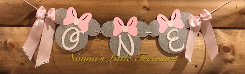 May include: A silver glitter banner with pink bows and the word "ONE" in white letters. The banner is decorated with pink and white Minnie Mouse ears. The text "Nonna's Little Treasure" is printed below the banner.