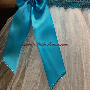 May include: A white tulle tutu with a turquoise satin bow and a row of turquoise rhinestones around the waist. The text "Nonna's Little Treasures" is visible on the tulle.