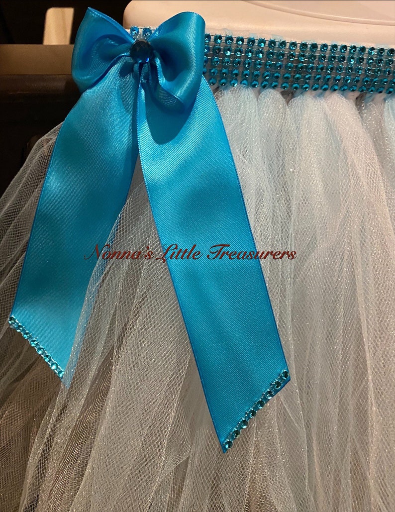 May include: A white tulle tutu with a turquoise satin bow and a row of turquoise rhinestones around the waist. The text "Nonna's Little Treasures" is visible on the tulle.
