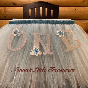 May include: A light blue tulle tutu with a silver glitter "ONE" banner and snowflake embellishments. The tutu is decorated with a silver and blue rhinestone trim. The banner reads "Nonna's Little Treasures".