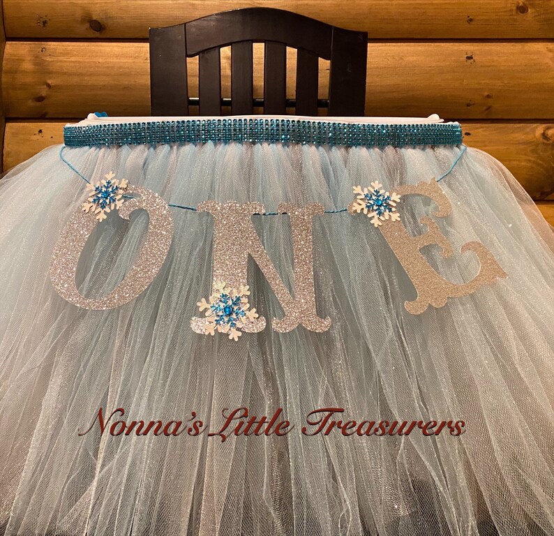May include: A light blue tulle tutu with a silver glitter "ONE" banner and snowflake embellishments. The tutu is decorated with a silver and blue rhinestone trim. The banner reads "Nonna's Little Treasures".