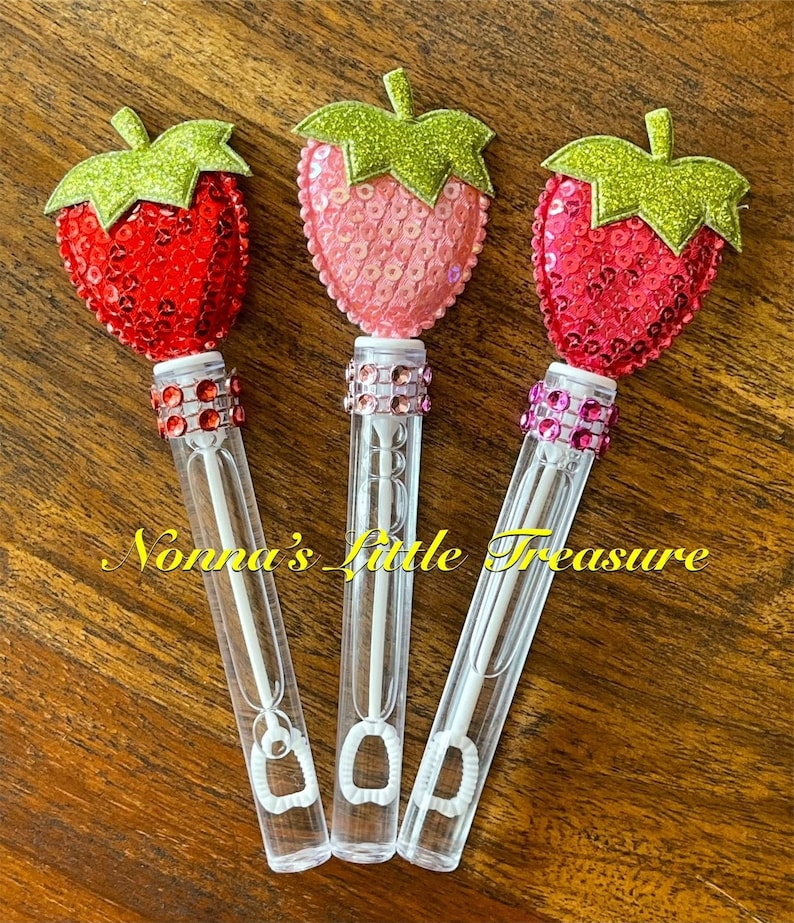 May include: Three clear plastic bubble wands with a strawberry-shaped topper. The strawberries are decorated with red, pink, and white sequins and green glitter. The wands are decorated with rhinestones.