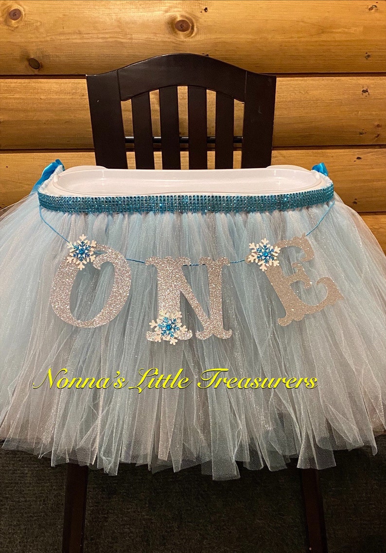 May include: A light blue tulle high chair skirt with a silver glitter "ONE" banner and blue snowflake embellishments. The skirt is decorated with a blue and silver sparkly trim. The text "Nonna's Little Treasures" is printed on the skirt.