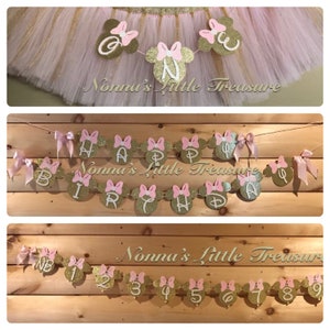 May include: A pink and gold Minnie Mouse themed birthday banner with the words "Happy Birthday" and a pink bow on each letter. The banner is strung on a white string and is hanging from a wooden surface. The banner is decorated with gold glitter.