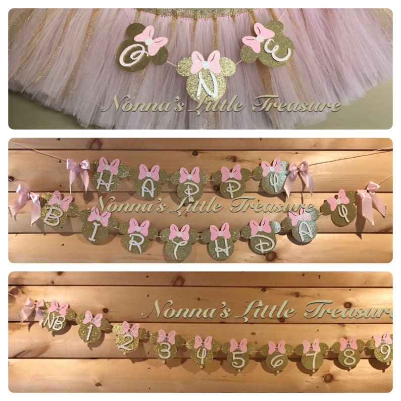 May include: A pink and gold Minnie Mouse themed birthday banner with the words "Happy Birthday" and a pink bow on each letter. The banner is strung on a white string and is hanging from a wooden surface. The banner is decorated with gold glitter.