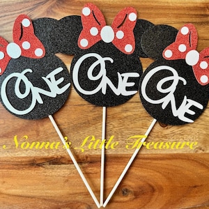 May include: Three black glittery mouse-shaped cake toppers, each featuring a red glittery bow with white polka dots and the word ONE in white script lettering. Each topper is attached to a thin white stick for placement in a cake or food item.