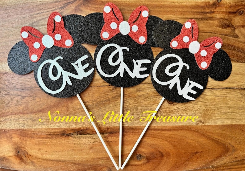 May include: Three black glittery mouse-shaped cake toppers, each featuring a red glittery bow with white polka dots and the word ONE in white script lettering. Each topper is attached to a thin white stick for placement in a cake or food item.