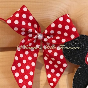May include: A red bow with white polka dots and a pearl-like embellishment. The bow is part of a larger design, with a black glittery silhouette of a character's head visible on the right. The text "@Nonnas Little Treasure" is also visible.