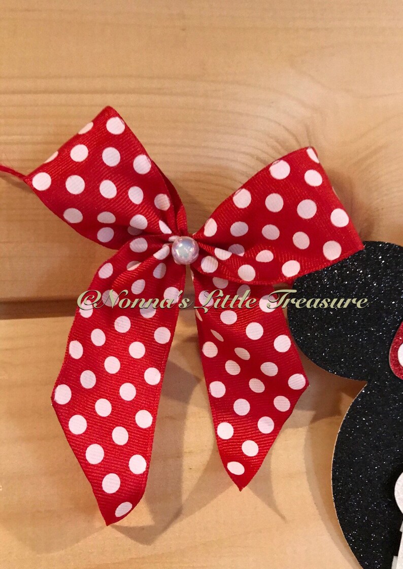 May include: A red bow with white polka dots and a pearl-like embellishment. The bow is part of a larger design, with a black glittery silhouette of a character's head visible on the right. The text "@Nonnas Little Treasure" is also visible.