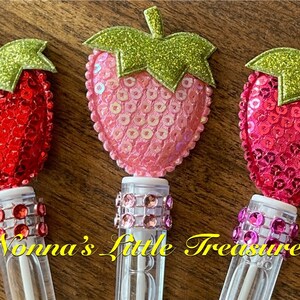 May include: Three clear plastic tubes with a white top and a sparkly, glittery strawberry on top. The strawberries are red, pink, and fuchsia. The tubes are decorated with a band of rhinestones.