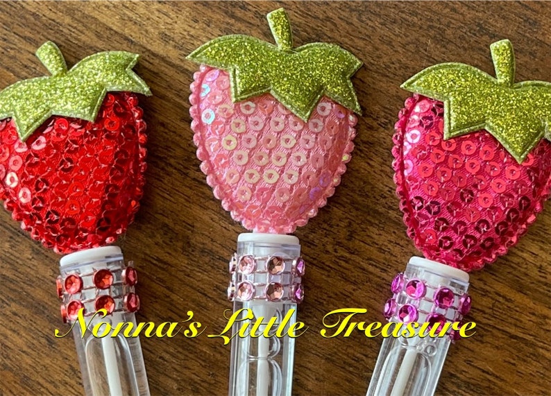 May include: Three clear plastic tubes with a white top and a sparkly, glittery strawberry on top. The strawberries are red, pink, and fuchsia. The tubes are decorated with a band of rhinestones.