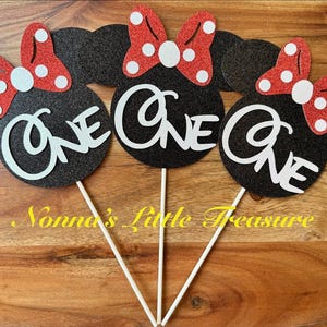 May include: Three black glittery mouse-shaped cake toppers on white sticks. Each topper features a red glittery bow with white polka dots and the word ONE in white script lettering. The design includes circular ears and a central bow detail.