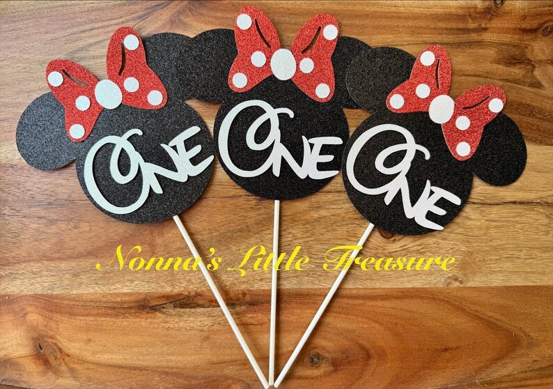 May include: Three black glittery mouse-shaped cake toppers on white sticks. Each topper features a red glittery bow with white polka dots and the word ONE in white script lettering. The design includes circular ears and a central bow detail.