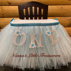 May include: A light blue tulle high chair skirt with a silver glitter "ONE" banner and snowflake embellishments. The skirt is decorated with a turquoise and silver ribbon and a turquoise rhinestone trim. The text "Nonna's Little Treasures" is printed on the skirt.