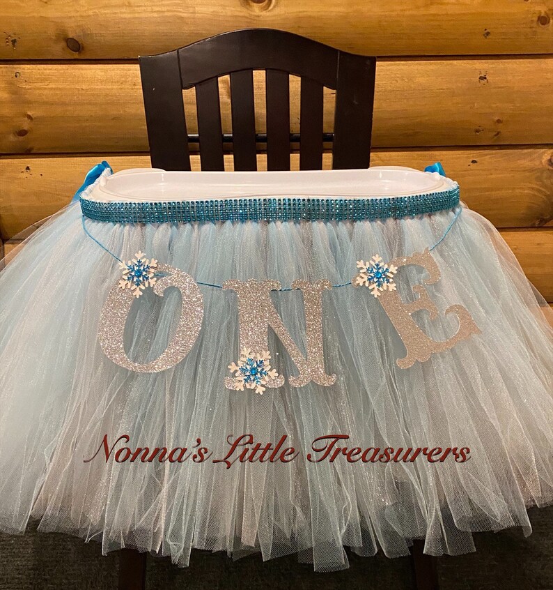 May include: A light blue tulle high chair skirt with a silver glitter "ONE" banner and snowflake embellishments. The skirt is decorated with a turquoise and silver ribbon and a turquoise rhinestone trim. The text "Nonna's Little Treasures" is printed on the skirt.
