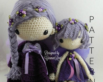 May include: Two crocheted dolls with purple and white yarn. The larger doll has a purple dress and a braid in her hair. The smaller doll has a purple dress and a headband with flowers. The dolls are sitting on a white surface. The text "Designed by Carmen Rent" is visible in the image.