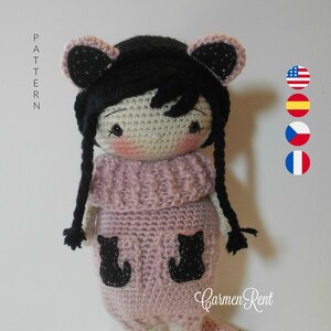 Op de afbeelding: Gehaakte pop met zwart haar, een roze trui en zwarte kattendetails. De pop heeft een staart en staat op een wit oppervlak. De tekst "Carmen Rent" is zichtbaar rechtsonder.