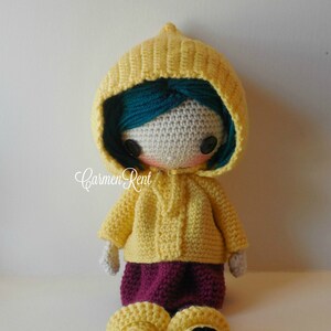 May include: A crocheted doll wearing a yellow raincoat with a hood, a burgundy skirt, and black boots. The doll has teal hair and black button eyes.