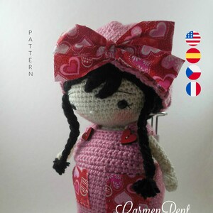 May include: A crocheted doll with pink overalls and a red heart-patterned bow. The doll has black hair and brown eyes. The doll is standing on a white base with the text "Carmen Rent" written on it.