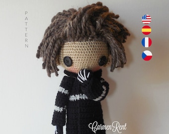 May include: A handmade crochet doll with brown yarn hair, black button eyes, and a tan face. The doll wears a black sweater with white stripes, light blue shorts, and brown shoes. The doll is standing on a silver stand. The text "Carmen Rent" is visible.