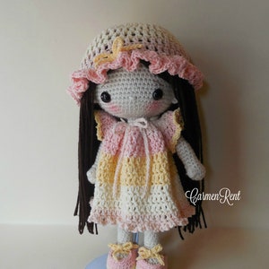 May include: A crocheted doll with long brown hair, wearing a pink and yellow dress, a pink and white hat, and pink and yellow shoes. The doll is standing on a blue stand. "Carmen Rent" is written in the bottom right corner of the image.