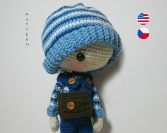 Könnte beinhalten: Eine gehäkelte Puppe mit einem blau-weiß gestreiften Hut, blauen Latzhosen und einer braunen Tasche. Die Puppe hat einen hellbraunen Teint und schwarze Augen. Die Puppe steht auf einer weißen Oberfläche.