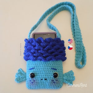 Op de afbeelding: Een gehaakte blauwe en turquoise telefoonhoes met een blauwe en witte gehaakte band. De hoes heeft een schattig cartoon gezicht met zwarte ogen en een kleine mond. De hoes is ontworpen om een smartphone te bevatten. De tekst "Carmen Rent" is zichtbaar in de rechterbenedenhoek van de afbeelding.