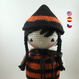 May include: A crocheted doll wearing a black and orange striped dress with a black ribbon. The doll has black hair, a black pointed hat with an orange stripe, and black shoes with orange accents.