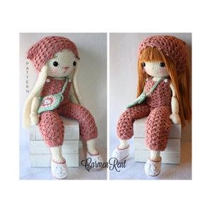 May include: Two crocheted dolls wearing pink overalls and white shoes. One doll has long brown hair and a pink crocheted hat. The other doll has white bunny ears and a pink crocheted hat. Both dolls are sitting on a white wooden box.