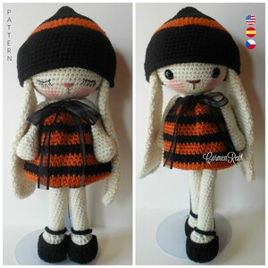 May include: A crocheted bunny doll wearing a black and orange striped dress and a black and orange striped hat. The doll has white fur and black eyes. The doll is standing on a white stand.
