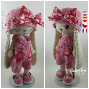 May include: A crocheted pink bunny with long white ears, wearing a pink hat with a red heart print bow, a pink overall with red heart print pockets, and pink shoes with red heart accents. The bunny is standing on a white platform.