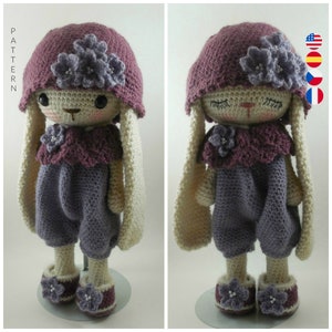 May include: A crocheted bunny doll wearing a purple hat and dress with white crocheted ears and shoes. The doll has a purple flower on its head and a purple flower on its dress.