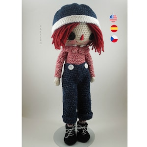 May include: Crocheted rag doll with red hair, a blue and white hat, a red and white striped shirt, blue jeans, and white buttons. The doll is standing on a white stand.