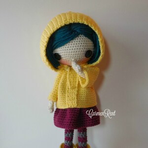 May include: Crocheted doll wearing a yellow raincoat, a burgundy skirt, grey and white striped tights, and yellow rain boots. The doll has teal hair and is holding her hand to her face.