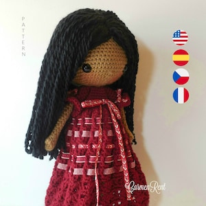 Könnte beinhalten: Häkelpuppe mit langen schwarzen Haaren, die ein rot-weiß kariertes Kleid mit einem roten Band trägt. Die Puppe hat braune Haut und große braune Augen.
