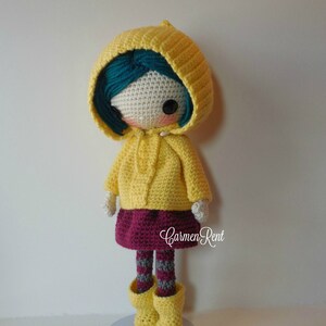 May include: A crocheted doll wearing a yellow raincoat, a burgundy skirt, and grey and white striped tights. The doll has teal hair and brown eyes. The doll is standing on a light blue base.
