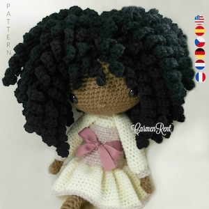 Puede incluir: Muñeca de crochet con piel marrón, cabello negro rizado y un vestido blanco con un lazo rosa. La muñeca lleva zapatos rosas con detalles florales. La muñeca está sentada sobre una superficie blanca. El texto "Carmen Rent" es visible en la imagen.