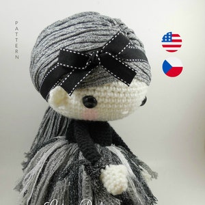 May include: A handmade crochet doll with a gray and black yarn hairstyle, adorned with a black bow. The doll wears a black top and a layered skirt in shades of gray and black. The doll's face is white with black button eyes and rosy cheeks.
