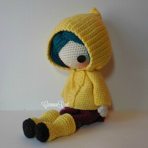 May include: A crocheted doll wearing a yellow raincoat with a hood, blue hair, and black boots. The doll is sitting on a white surface.