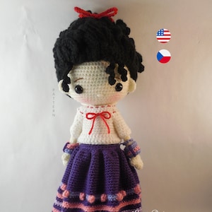 May include: Crocheted doll with black curly hair, a white shirt with a red bow, a purple skirt with pink and purple stripes, and red shoes. The doll is standing on a white surface. The text "Carmen Rent" is visible in the bottom right corner of the image.